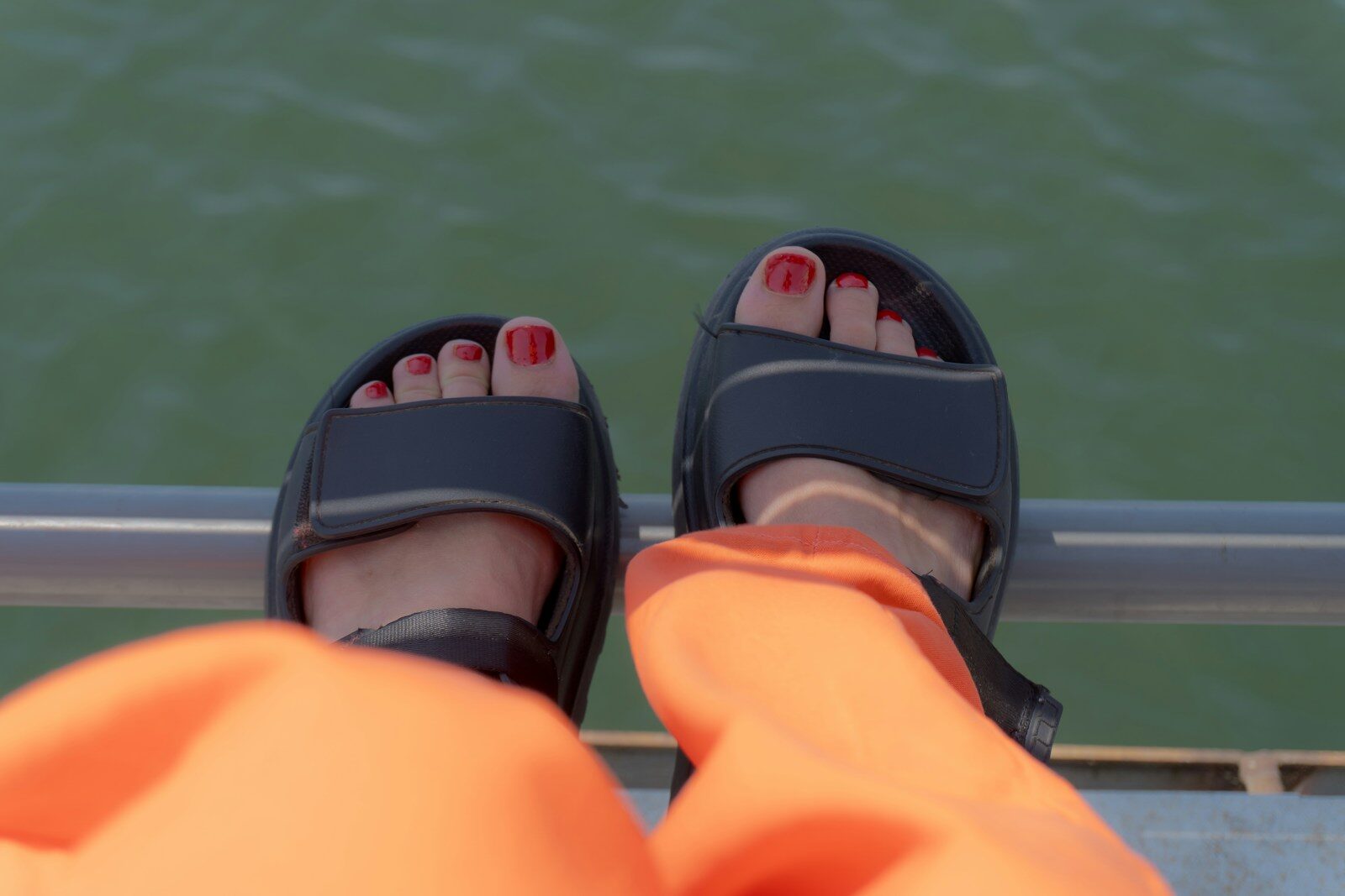Feet in sandals with red nail polish near water.